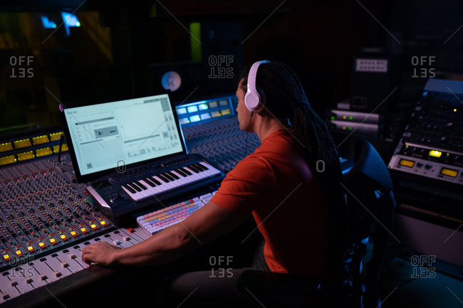 Side view of a young mixed race male sound engineer sitting and working at a mixing desk in a recording studio using a computer and wearing headphones