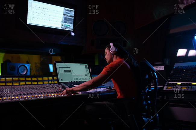 Side view of a young mixed race male sound engineer sitting and working at a mixing desk in a recording studio using a computer and wearing headphones