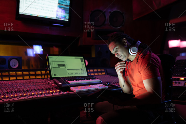 Side view close up of a young mixed race male sound engineer sitting and working at a mixing desk using a laptop computer in a recording studio