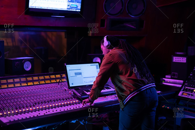Side view close up of a young mixed race male sound engineer standing at a mixing desk in a recording studio using a computer and wearing headphones