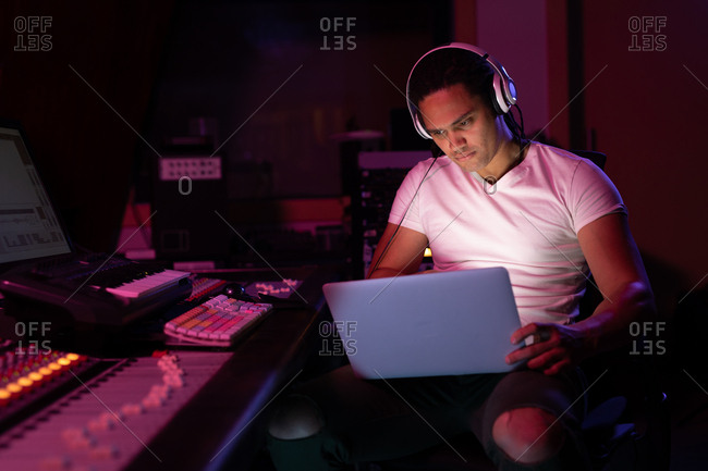 Front view close up of a young mixed race male sound engineer sitting and working at a mixing desk in a recording studio using a laptop and wearing headphones