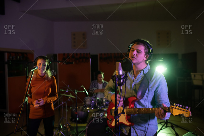 Front view of a young Caucasian female singer, a young Caucasian male singer guitarist and drummer performing during a session at a recording studio