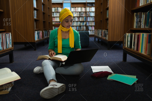 Front view close up of a young Asian female student wearing a hijab holding a book, using a laptop computer and studying in a library