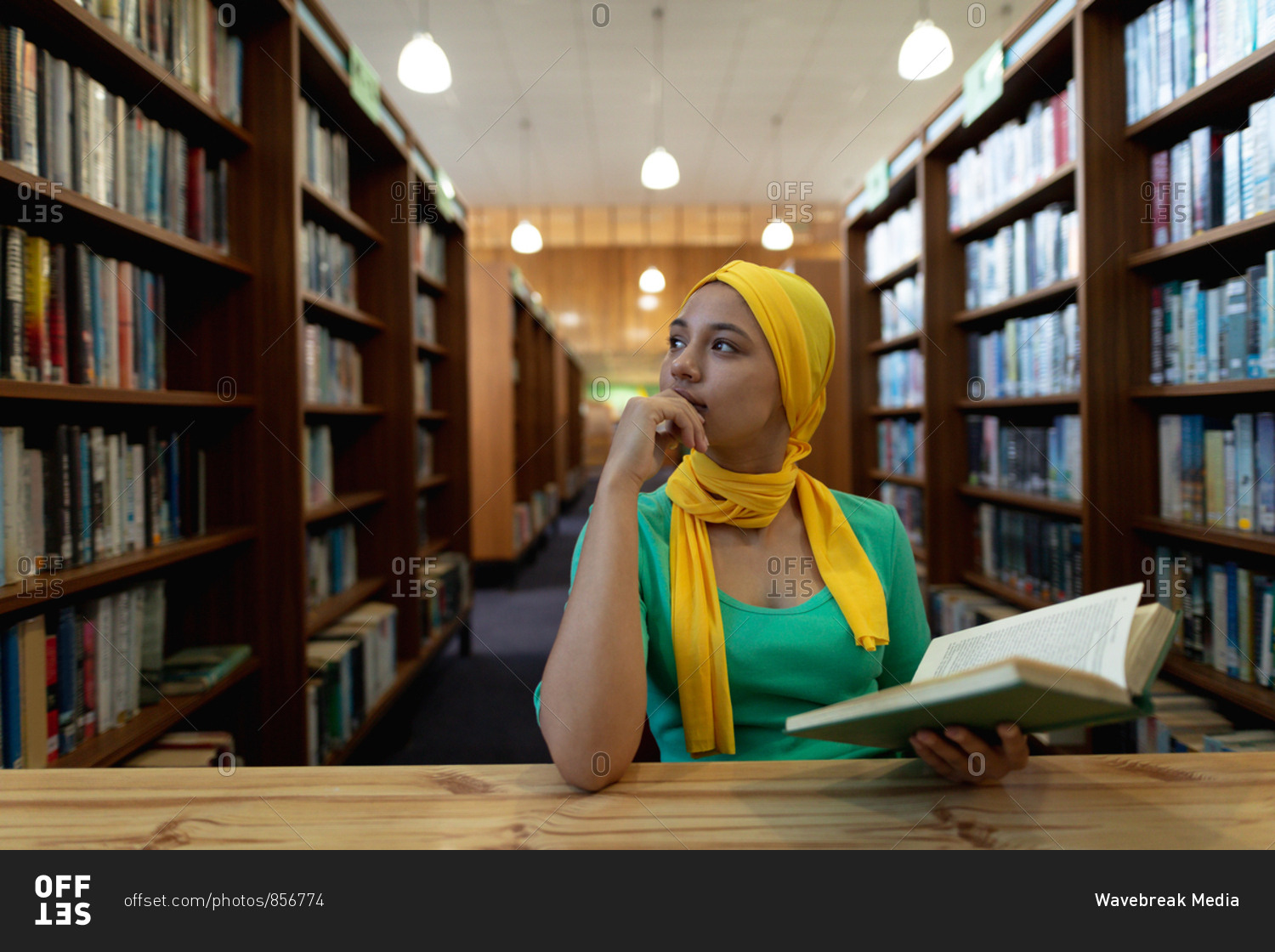 Front view close up of a young Asian female student wearing a hijab holding a book and studying in a library