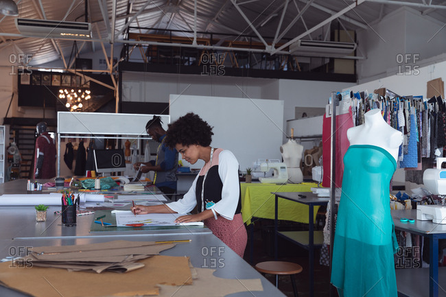 Side view of a young mixed race female fashion student working on a design in a studio at fashion college, with a mannequin withe dress on in the foreground and a male student working in the background