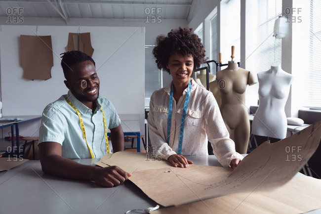 Front view of a young African American male and a young mixed race female fashion student holding a pattern while working on a design in a studio at fashion college, with mannequins in the background