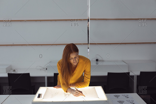 Front view of a young Caucasian female fashion student working on a design drawing on a lightbox in a studio at fashion college