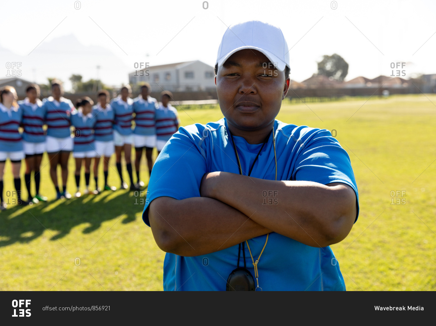 Portrait close up of a middle aged mixed race female rugby coach standing on a rugby pitch with her arms crossed looking to camera, with her team standing in a row in the background
