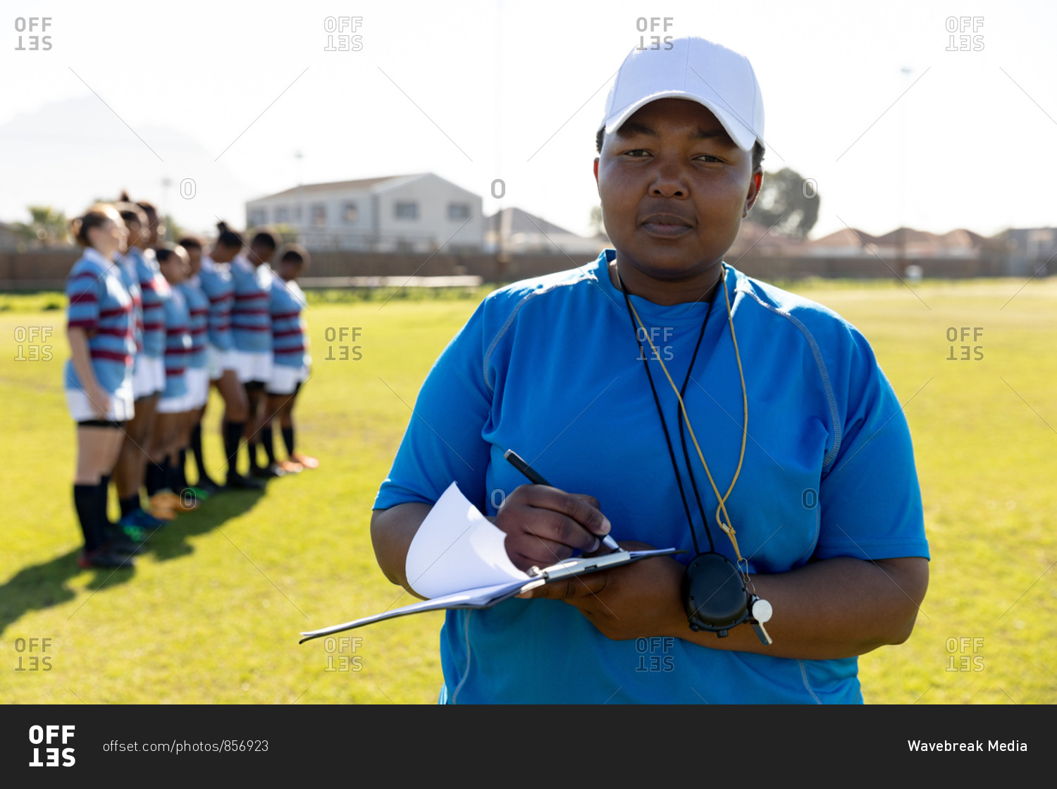 Front view close up of a middle aged mixed race female rugby coach standing on a rugby pitch writing on a clipboard and looking to camera, with her team standing in a row together in the background