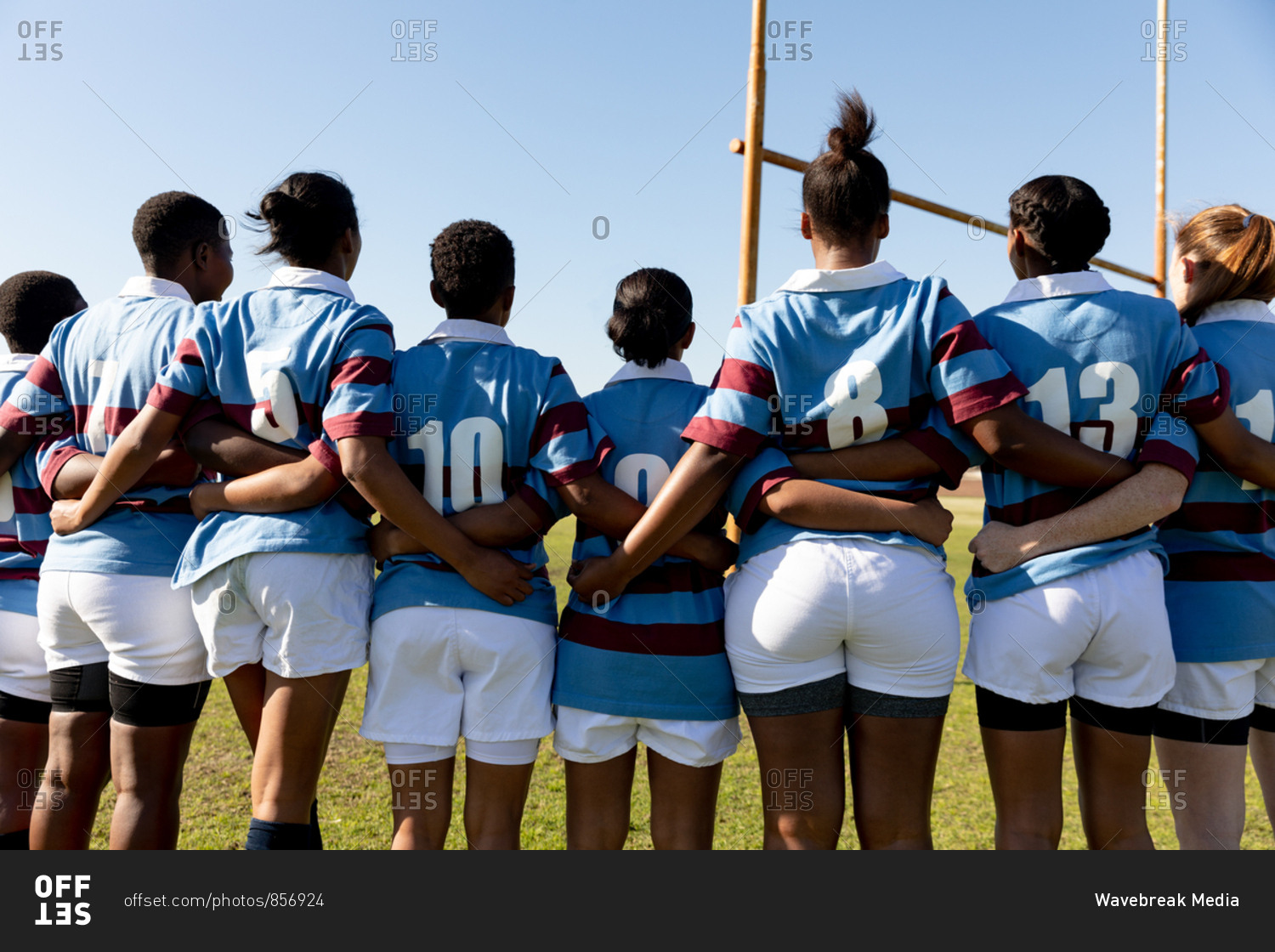 Rear view of a team of young adult multi-ethnic female rugby players standing on a rugby pitch with arms linked preparing for a rugby match