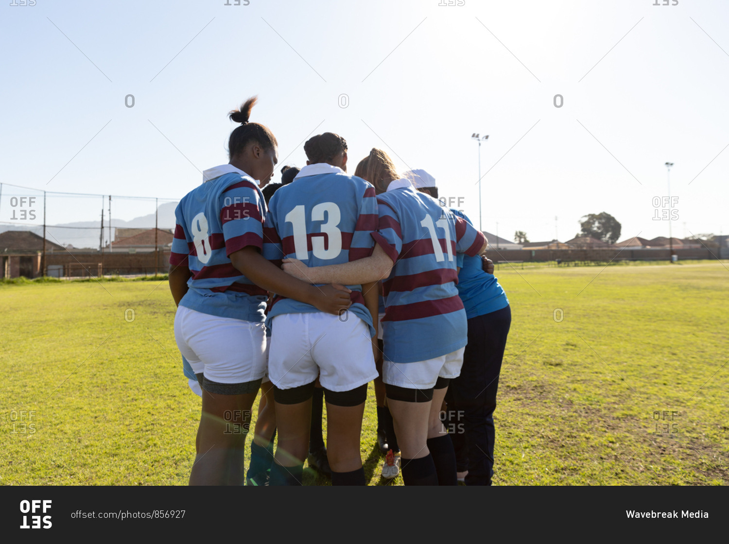 Rear view of a team of young adult multi-ethnic female rugby players and their coach standing in a huddle on a rugby field preparing for a rugby match