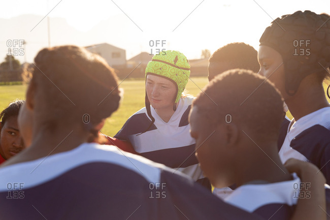Over the shoulder view of a team of young adult multi-ethnic female rugby players standing on a rugby field with arms linked preparing for a rugby match