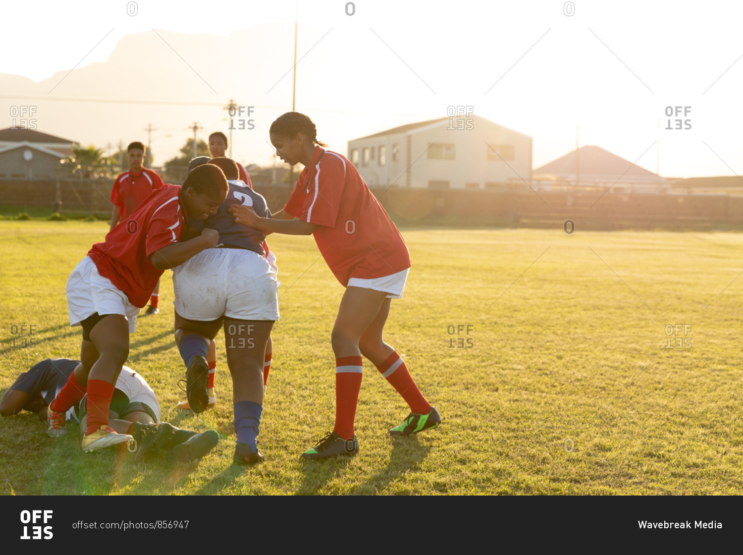 Side view of two young adult mixed race female rugby players tackling a player from the opposing team during a rugby match, with other players in the background and one lying on the ground