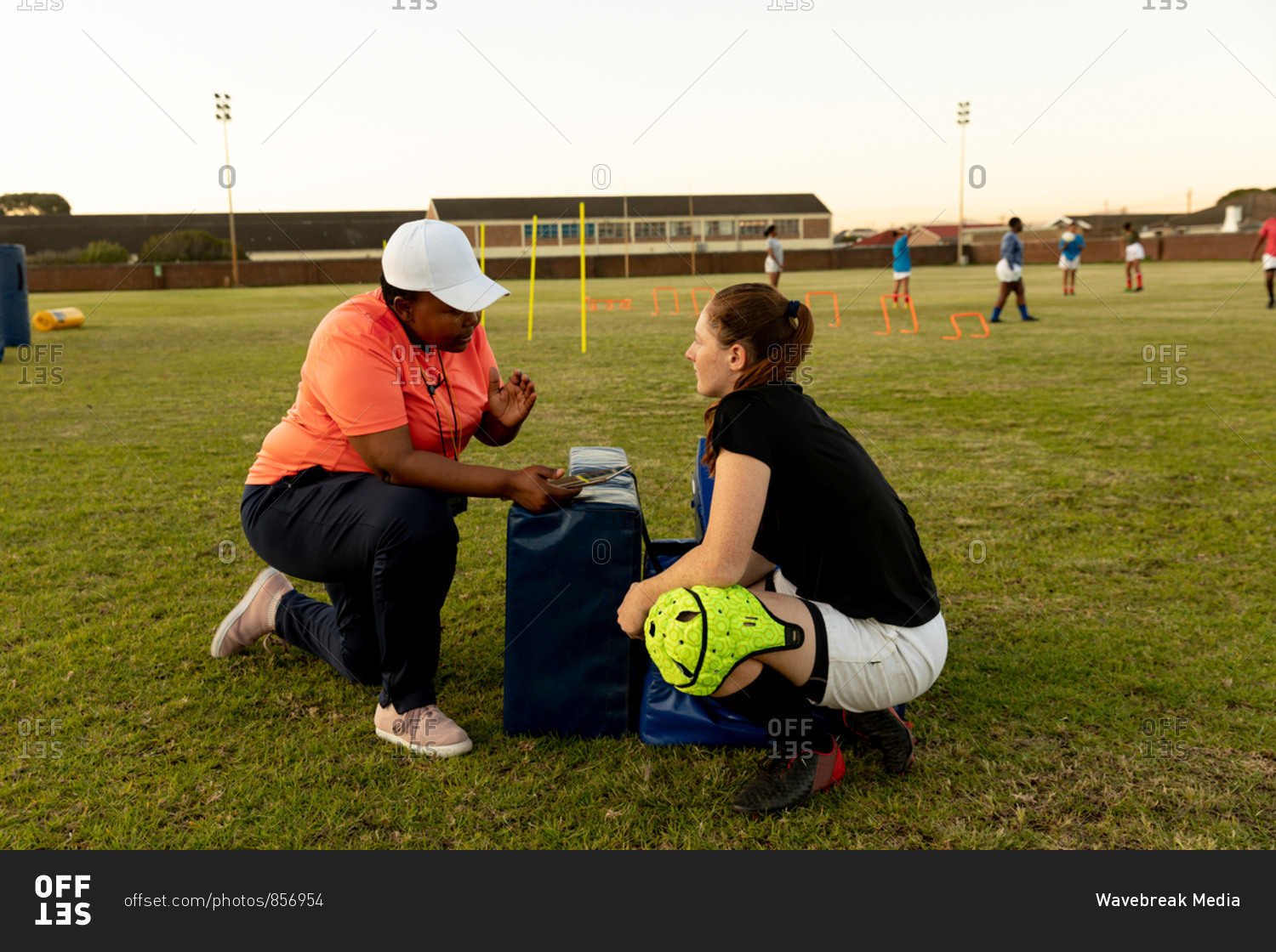 Side view of a middle aged mixed race female rugby coach kneeling and talking to a young adult Caucasian female rugby player on a rugby field during a training session