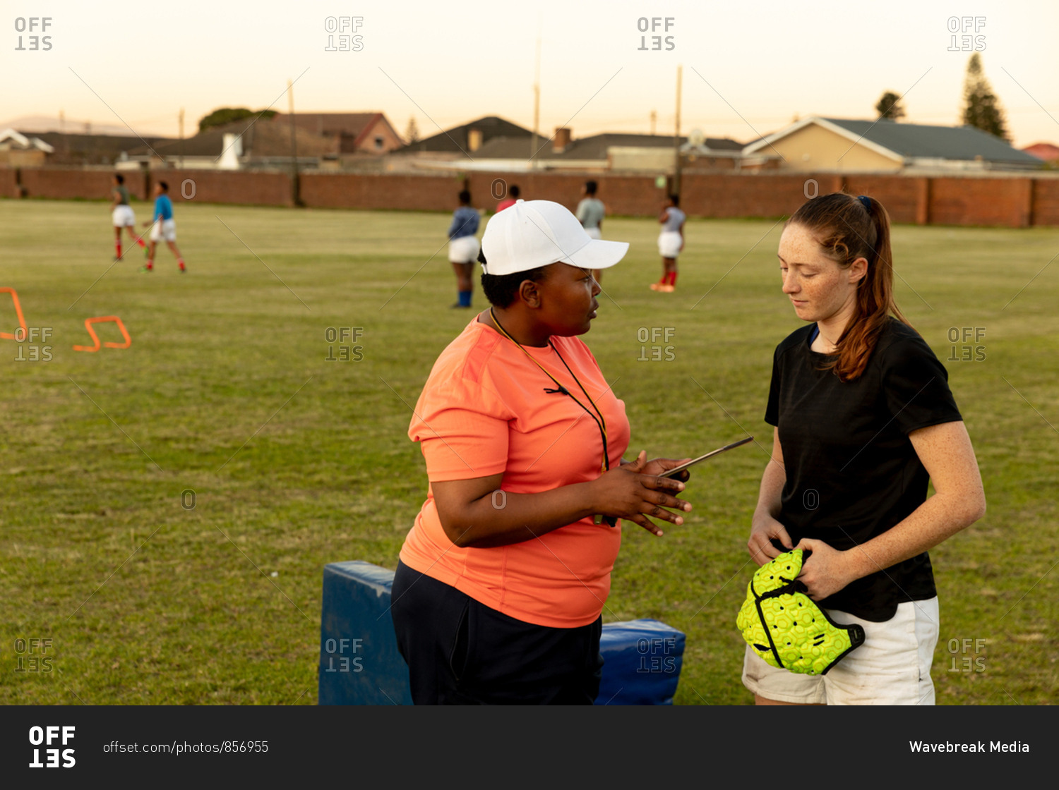 Side view of a middle aged mixed race female rugby coach talking to a young adult Caucasian female rugby player at a sports field during a training session