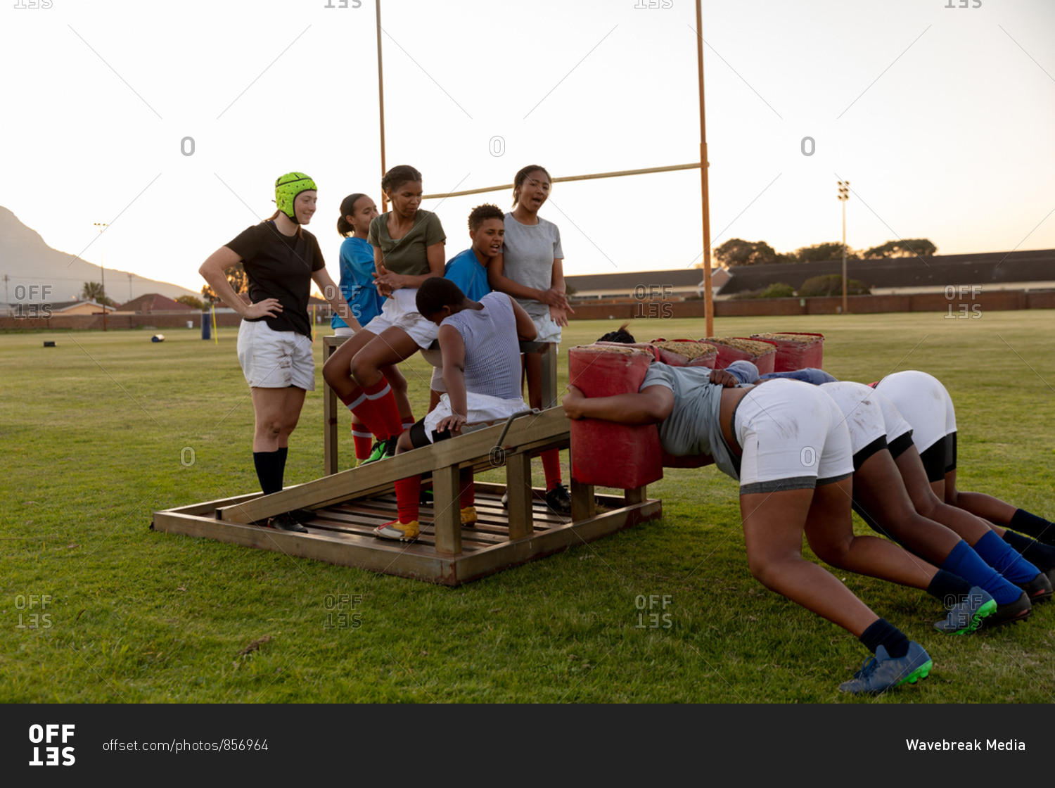 Side view of a team of a young adult multi-ethnic female rugby players on a sports field at a training session, with three of the young women practicing with a scrum machine while the others watch