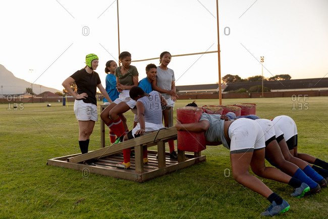 Side view of a team of a young adult multi-ethnic female rugby players on a sports field at a training session, with three of the young women practicing with a scrum machine while the others watch