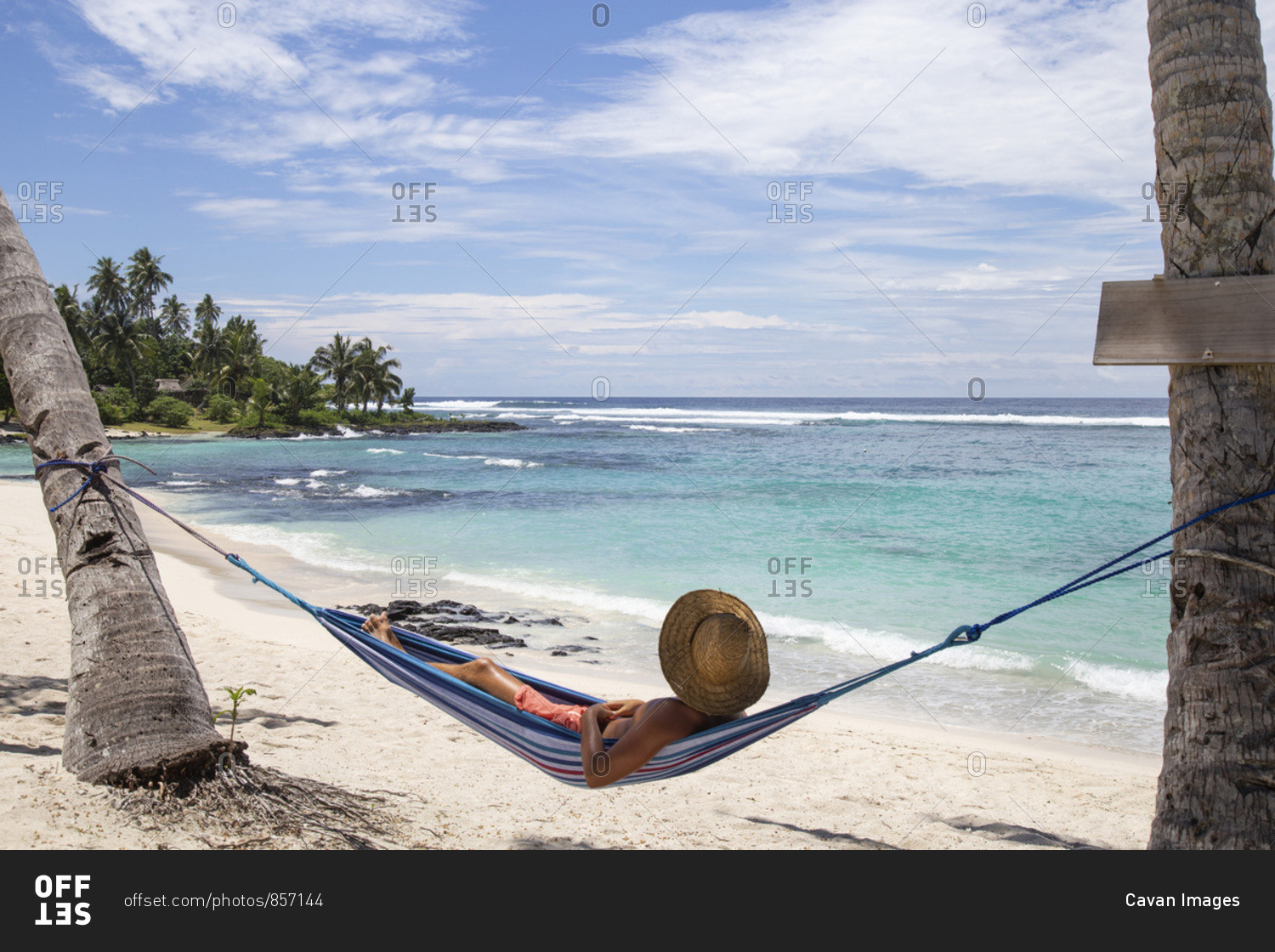 Man with hat, laying on blue hammock under palm trees at the beach