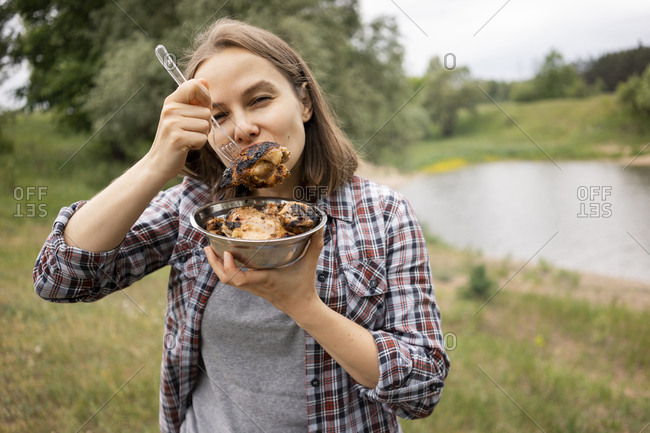 Woman standing at river bank and eating hot grilled chicken from bowl