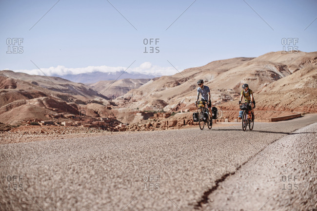 Morocco, Draa-Tafilalet, Telouet - April 8, 2019: Two bike packers ride along a road with desert mountains behind them