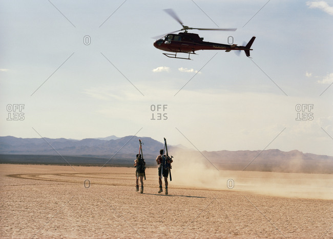 Adventurous young couple carrying sports equipment on their backs underneath hovering helicopter while running across a desert.