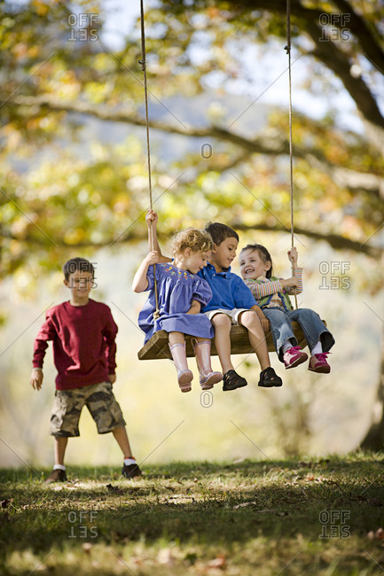 Three young siblings having fun on a swing together while their older brother pushes them in a grassy field.