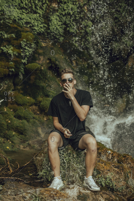 Portrait of a cool young man at a waterfall smoking a joint of marijuana