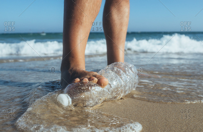 Foot of man stepping on empty plastic bottle lying on the beach- close-up