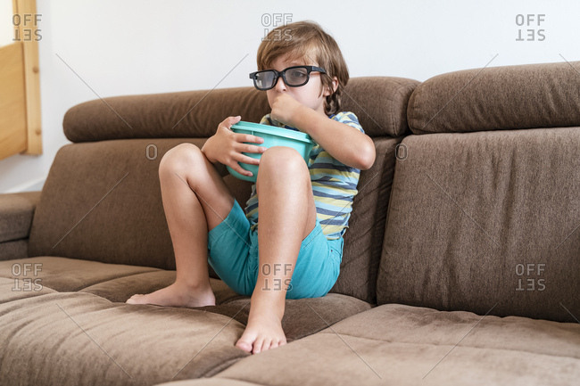 Boy sitting on couch at home wearing 3d glasses and eating popcorn