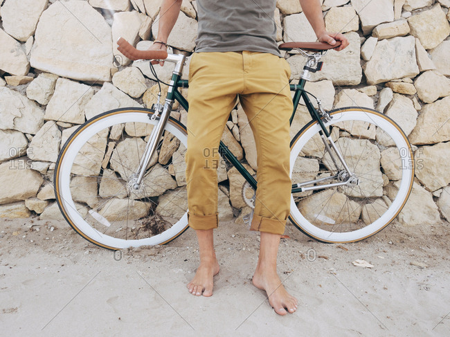 Barefoot man with Fixie bike in front of natural stone wall on the beach- partial view
