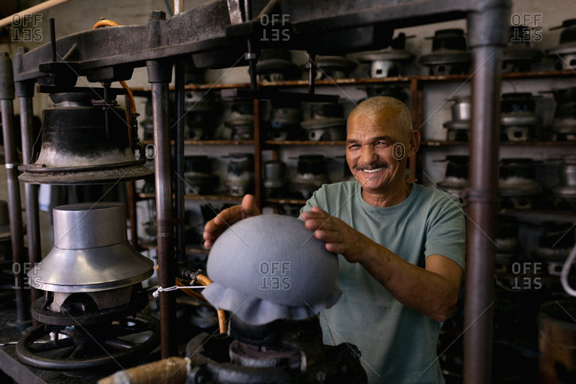 Portrait of a smiling senior mixed race man holding his hands above the top of a hat that has been shaped on a piece of equipment in the workshop at a hat factory
