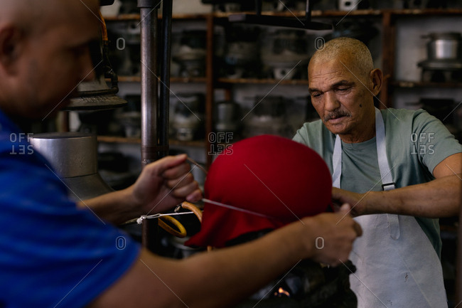 Side view of a senior and a middle aged mixed race man working together at a machine steaming the top of a hat to shape it in the workshop at a hat factory