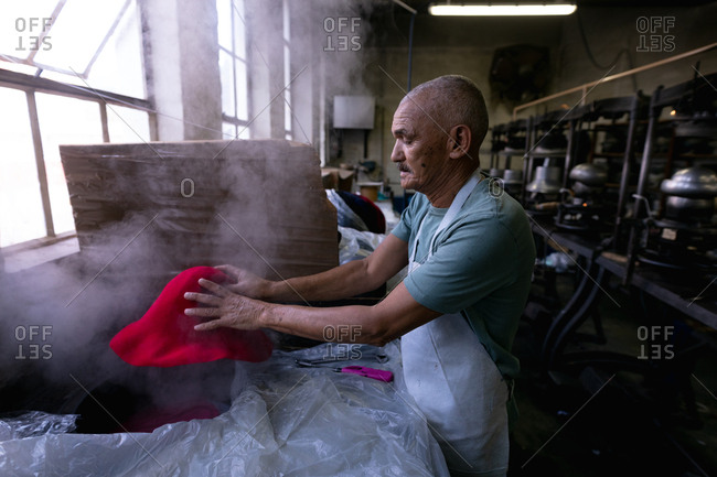 Side view of a senior mixed race man wearing an apron holding and shaping the top of a hat that has been steamed in the workshop at a hat factory