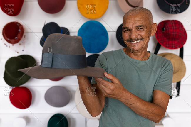 Portrait close up of a senior mixed race man smiling and holding a finished hat in the showroom at a hat factory