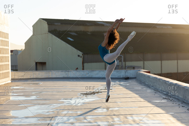 Side view of a young mixed race female ballet dancer jumping with her arms and one leg raised, on the rooftop of an urban building