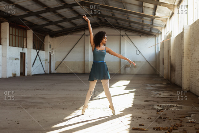 Front view of a young mixed race female ballet dancer standing on her toes with arms raised in a shaft of sunlight while dancing in an empty room at an abandoned warehouse