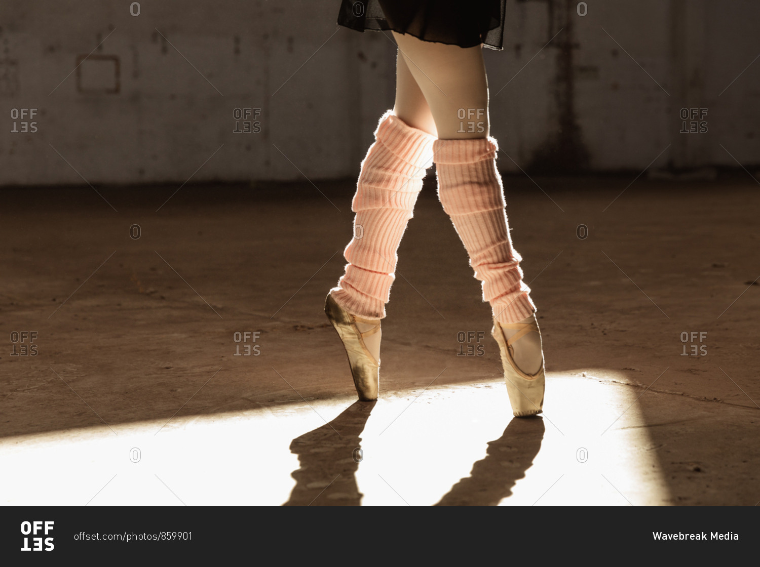 Low section of a young mixed race female ballet dancer wearing leg warmers and pointe shoes standing on her toes in shaft of sunlight while dancing in an empty room at an abandoned warehouse