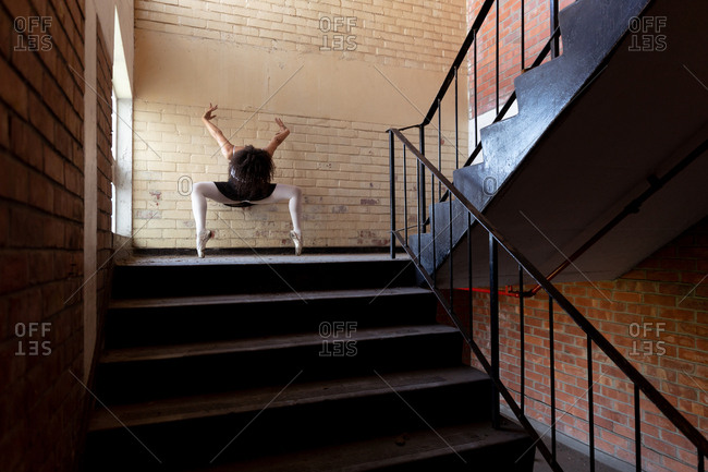 Front view of a young mixed race female ballet dancer holding a dance pose on her toes with arms raised and head down on a staircase landing in an abandoned warehouse