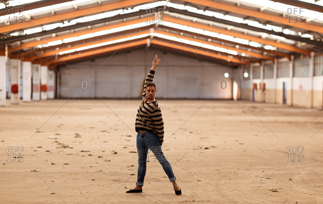 Front view of a young Caucasian male ballet dancer holding a dance pose with arm raised standing in an abandoned warehouse looking to camera