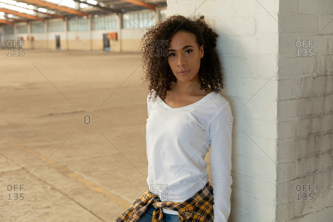Front view close up of a young mixed race woman with shoulder length curly hair and a shirt tied around her waist leaning against a pillar and looking to camera in an abandoned warehouse