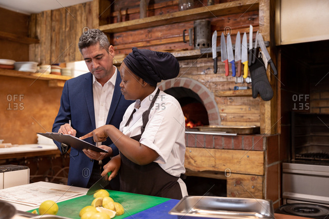Front view close up of a middle aged Caucasian restaurant manager holding a clipboard and talking with a young African American female chef in a restaurant kitchen