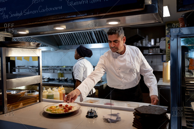 Front view close up of a middle aged Caucasian male chef putting out a dish ready for serving in a restaurant kitchen, while a female member of kitchen staff walks past behind him