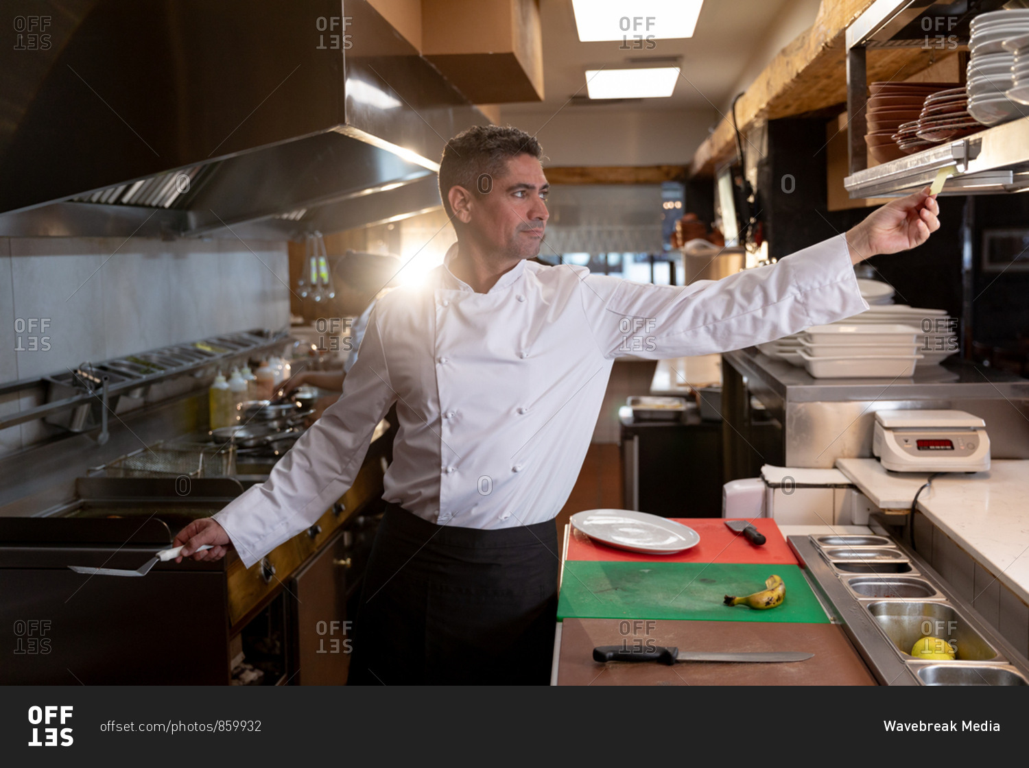 Front view close up of a middle aged Caucasian male chef reaching out to take an order at the order station in a restaurant kitchen
