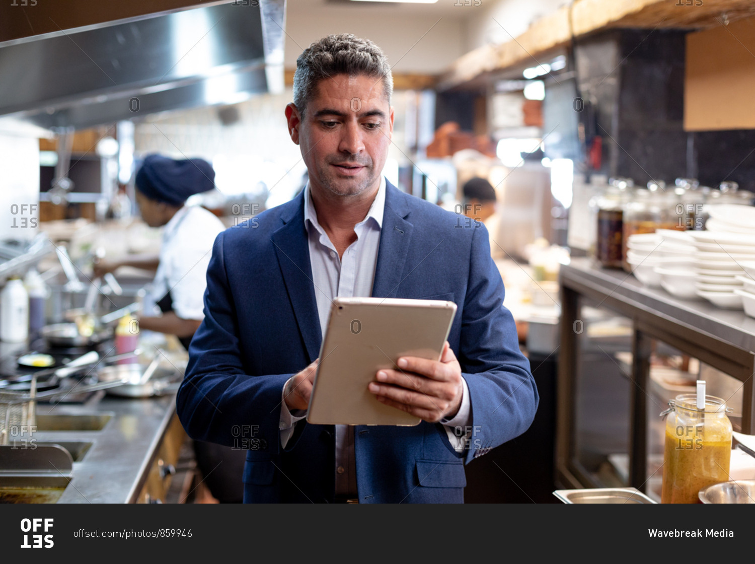 Front view close up of a middle aged Caucasian male restaurant manager using a tablet computer in a busy restaurant kitchen, while kitchen staff work in the background