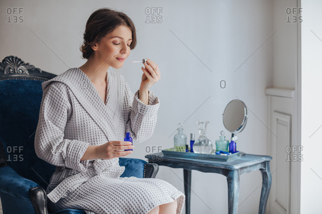 Beautiful Caucasian woman in bathrobe holding cosmetic serum bottle and smelling pipette.