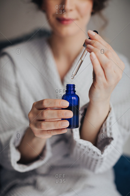Hands of unrecognizable woman in bathrobe holding bottle of cosmetic serum and a pipette.