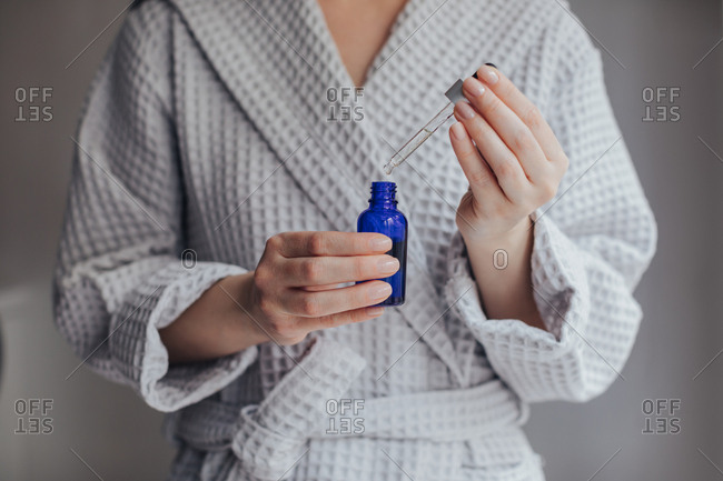 Hands of unrecognizable woman in bathrobe holding bottle of cosmetic serum and a pipette.
