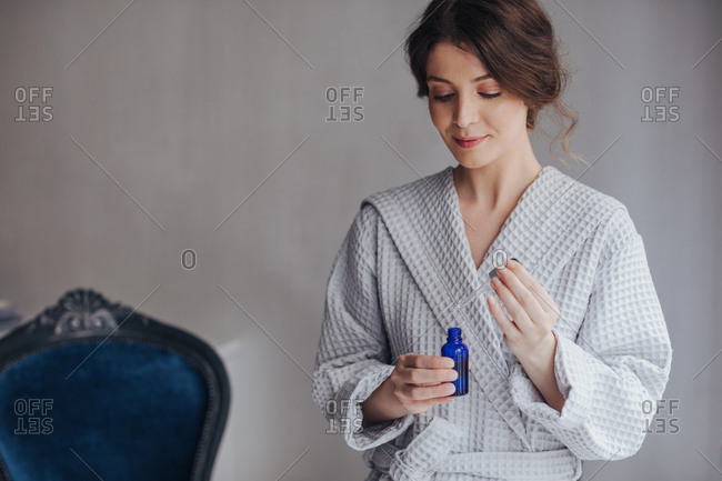 Beautiful Caucasian young woman in bathrobe holding cosmetic serum bottle and pipette.