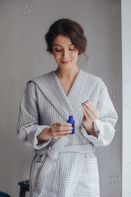 Beautiful Caucasian young woman in bathrobe holding cosmetic serum bottle and pipette.