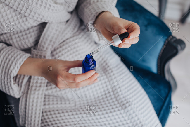 Hands of unrecognizable woman in bathrobe holding bottle of cosmetic serum and a pipette.